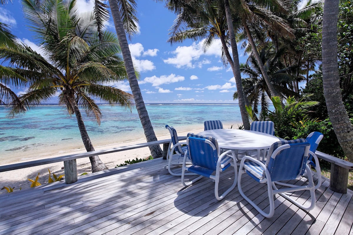 Una terraza de madera con una mesa y sillas, con vista a una playa de aguas turquesas cristalinas y palmeras.