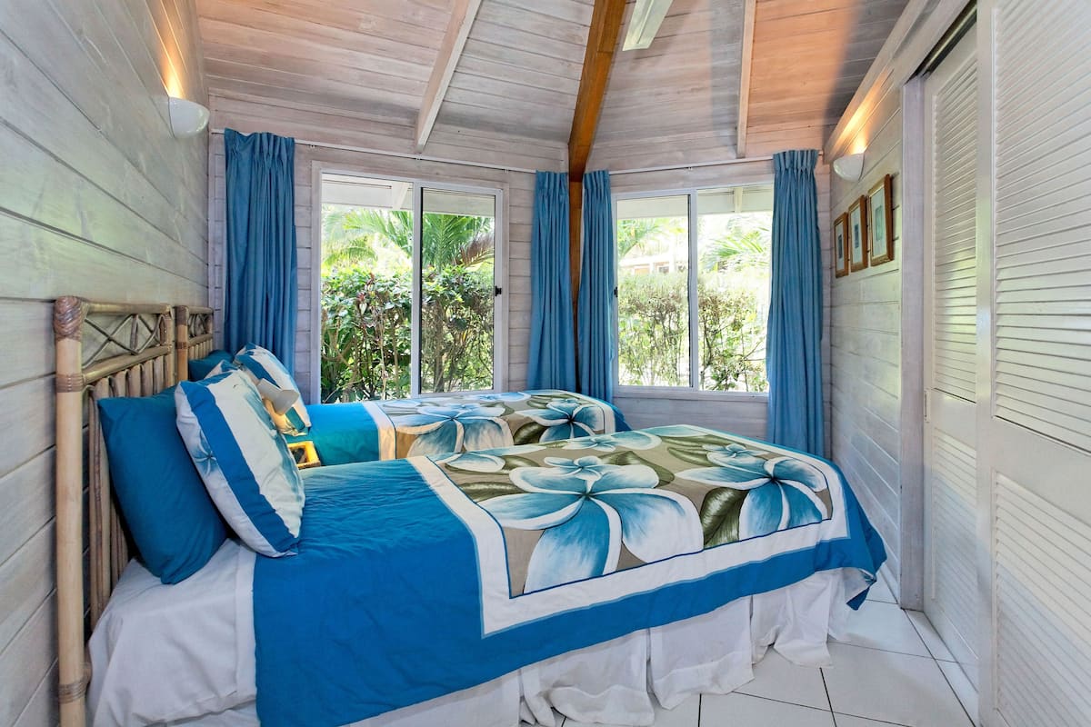 Un dormitorio con techo de madera, dos camas con ropa de cama azul y blanca, y ventanas grandes con cortinas azules.