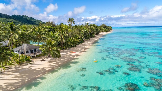 Una playa con agua turquesa cristalina, palmeras y una pequeña casa con techo de paja.