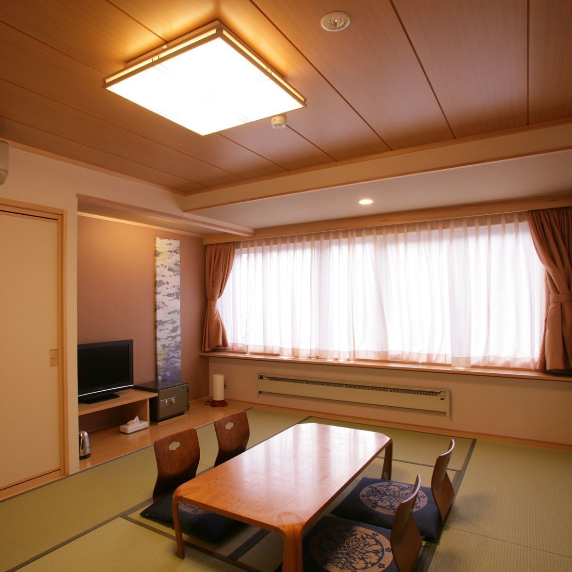 A hotel room with a wooden ceiling, a flat-screen TV, a dining table with chairs, a large window with curtains, and a wall-mounted air conditioner.