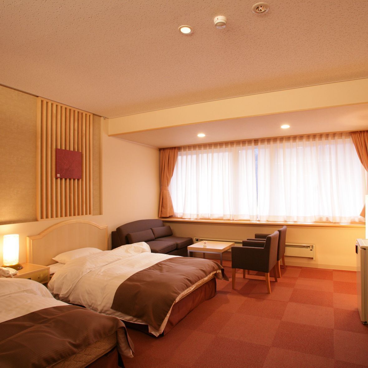 Habitación de hotel con dos camas, un escritorio, una silla, una lámpara, un radiador y una ventana con cortinas.