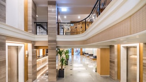Un hall d’hôtel spacieux, à plusieurs niveaux, avec un escalier central, des ascenseurs et un espace d’accueil.