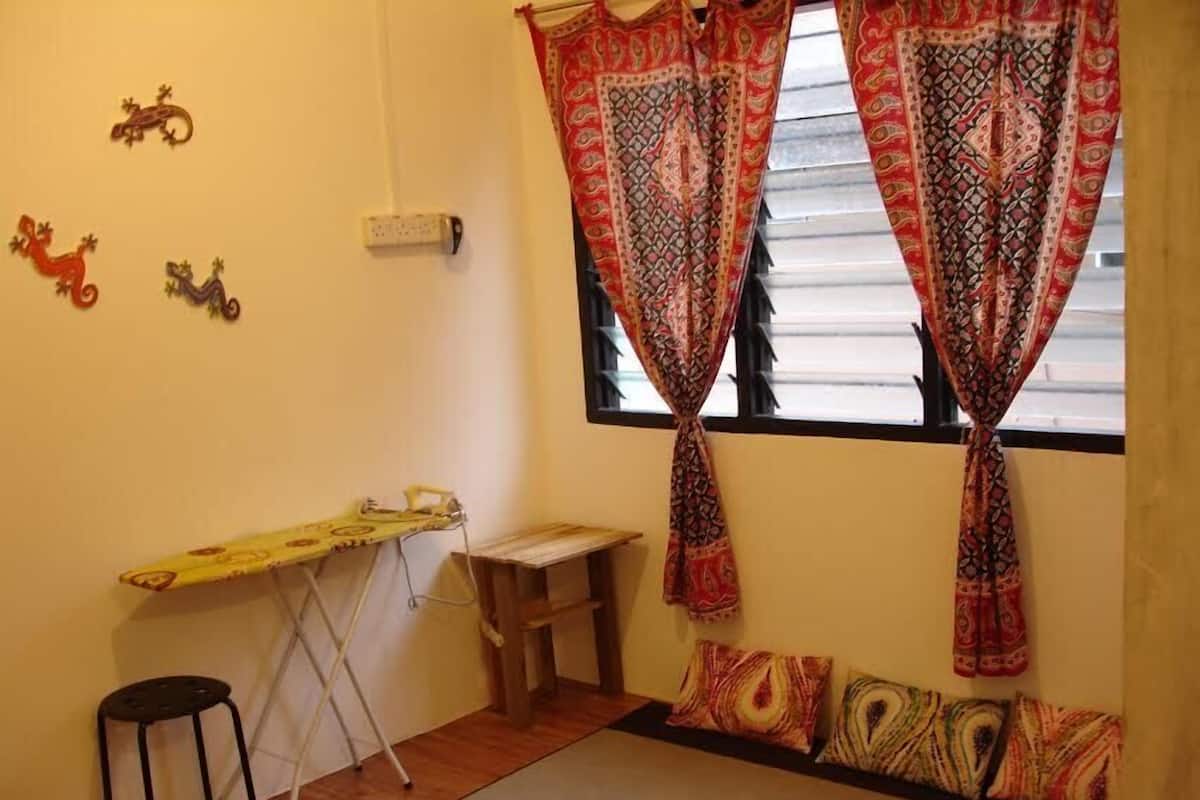 A small room with a window, a desk with an ironing board, a stool, and decorative pillows.