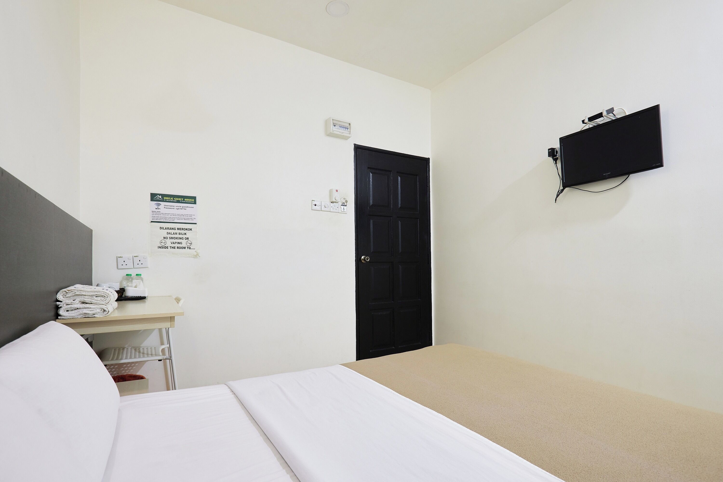 A hotel room with a bed, a desk with a computer, a TV mounted on the wall, and a door.