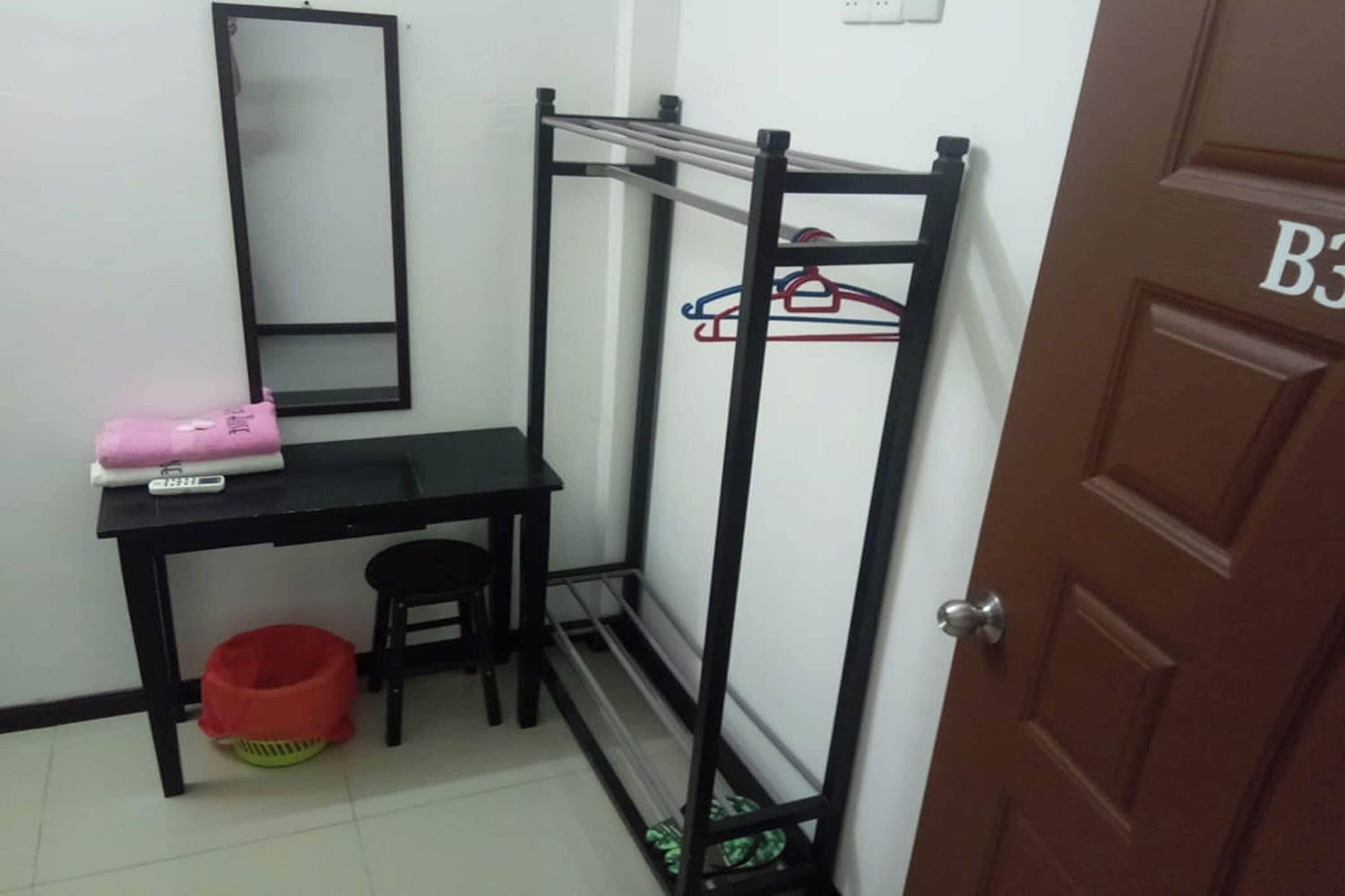 A small room with a black desk, a chair, a red basket, a black metal rack with clothes, and a brown door with a number.