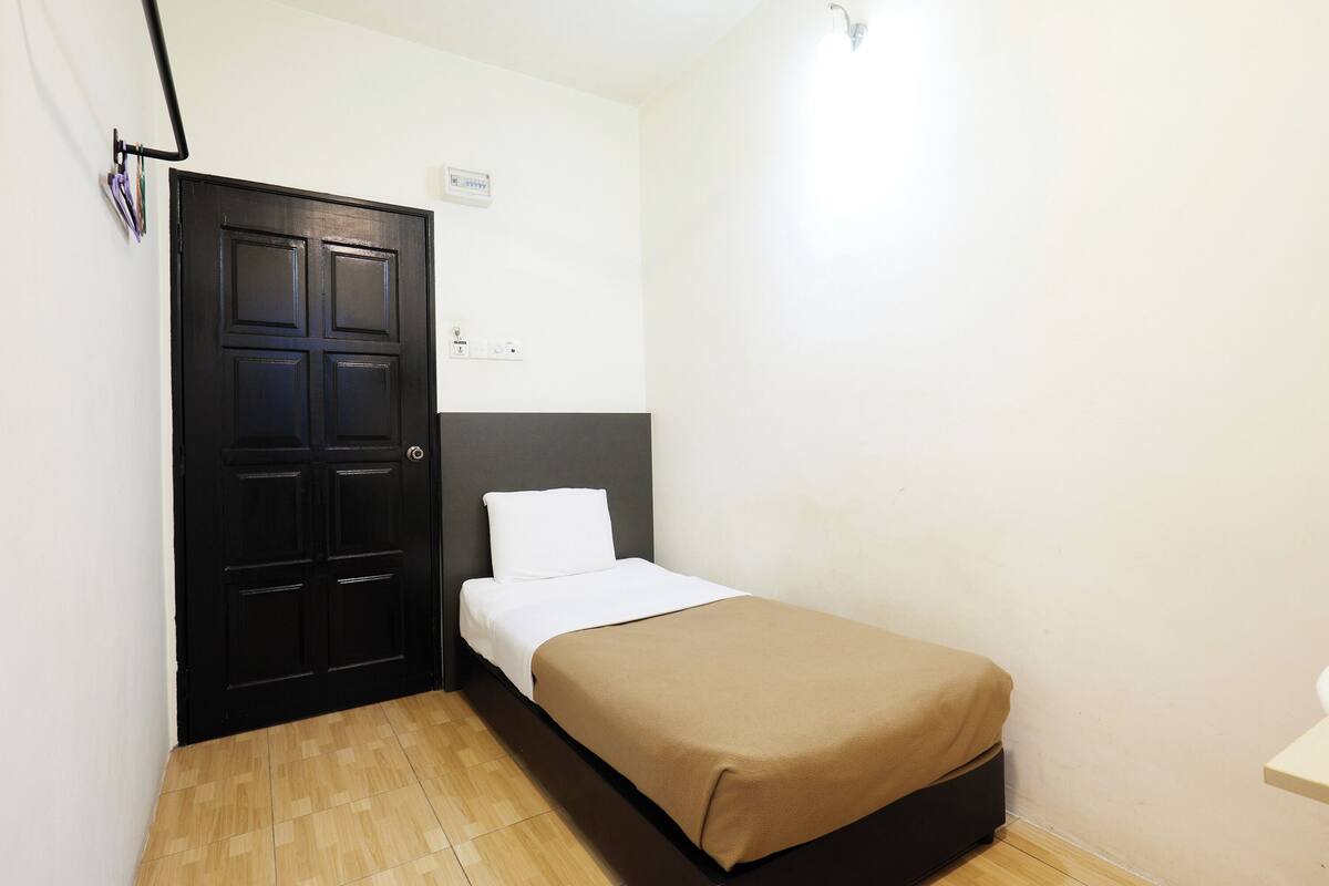Een kleine kamer met een enkel bed, een donker hoofdbord, een houten deur en een lichtgekleurde vloer.
