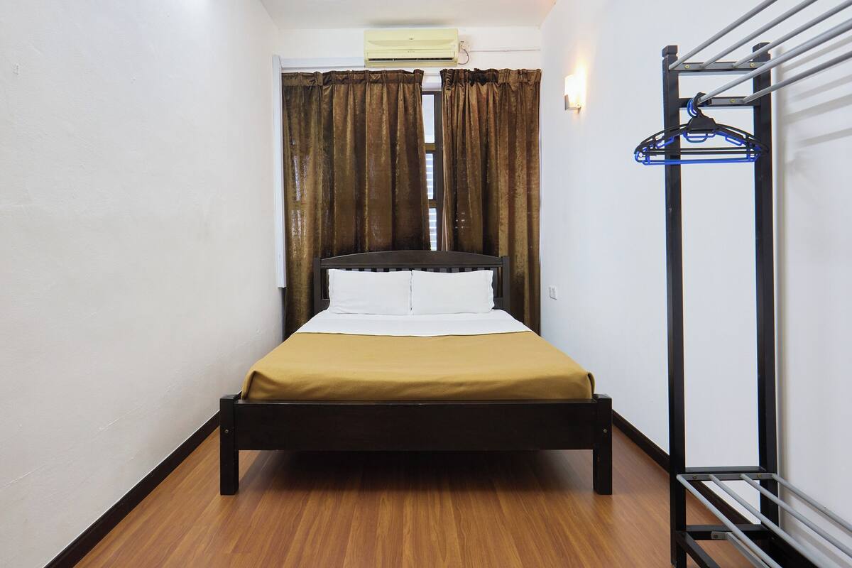 Een enkel bed met houten frame, een witte matras en een mosterdgele sprei. Er is een raam met bruine gordijnen, een airconditioning erboven en een metalen rek met kleding aan de rechterkant.