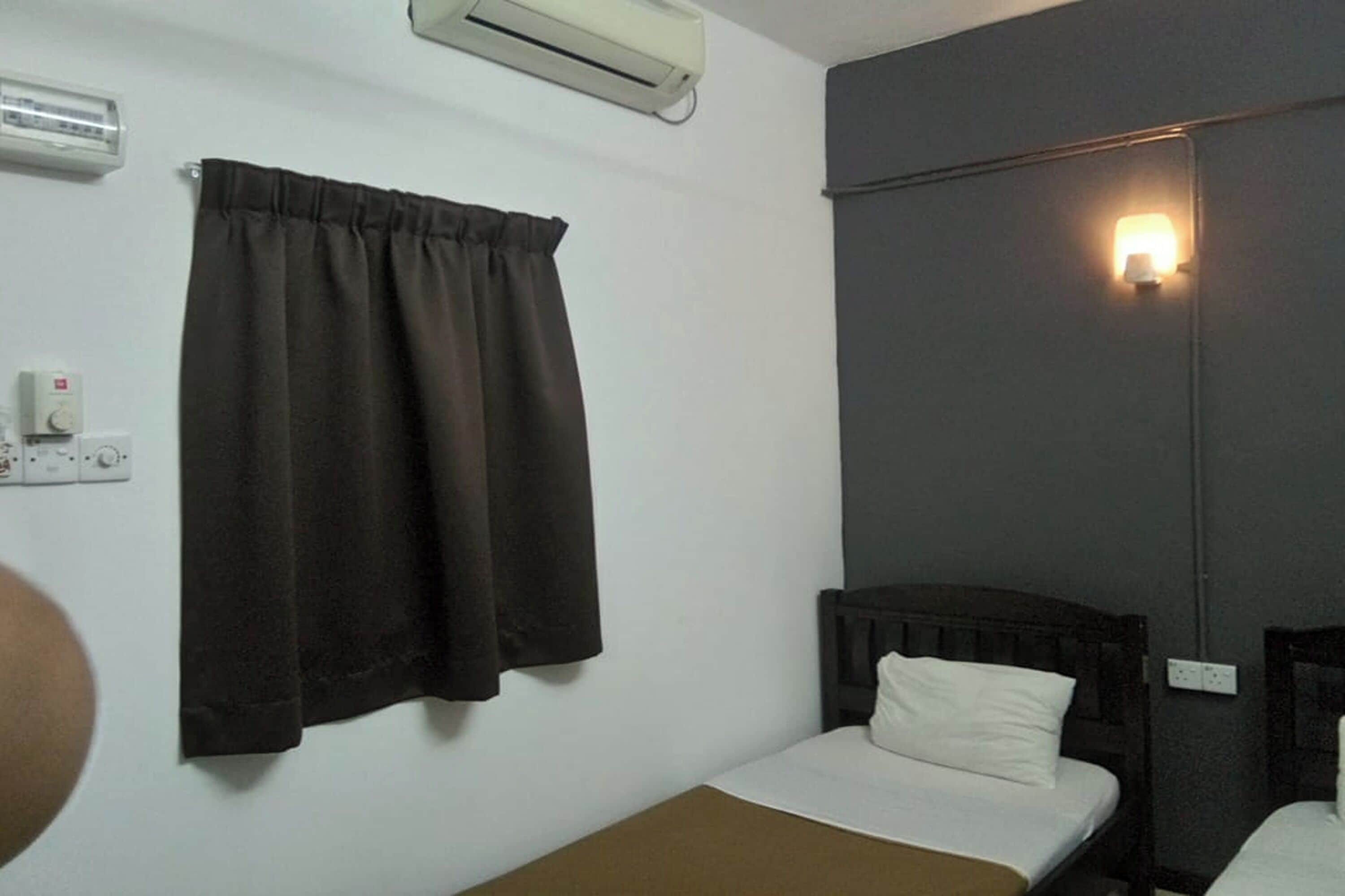 Een kleine eenpersoonskamer met een donker hoofdeinde, een bedlampje en een wandairconditioner.
