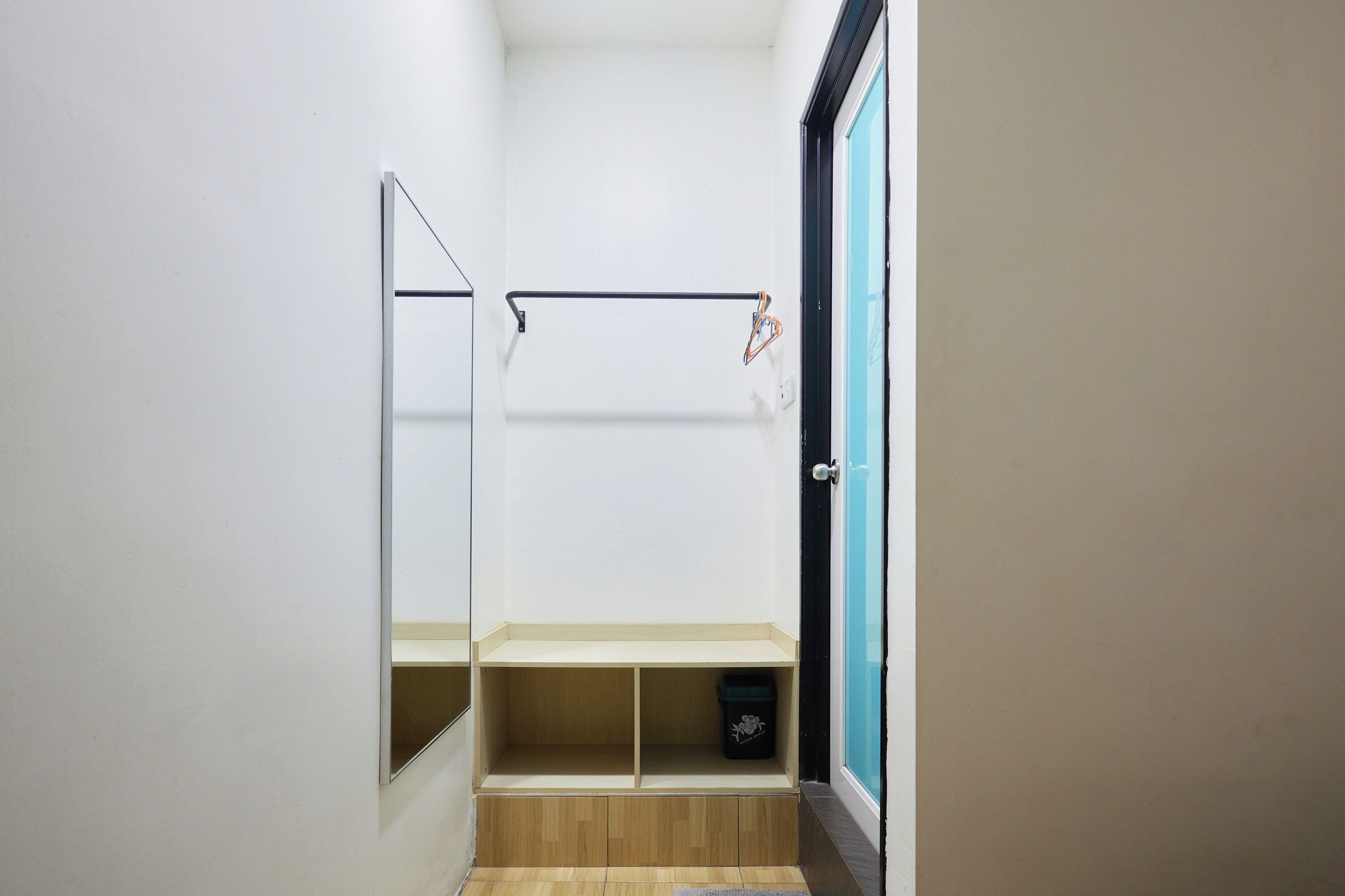 Une petite pièce étroite, avec un plancher en bois, un miroir, une porte noire et une étagère sur laquelle se trouve une poubelle.