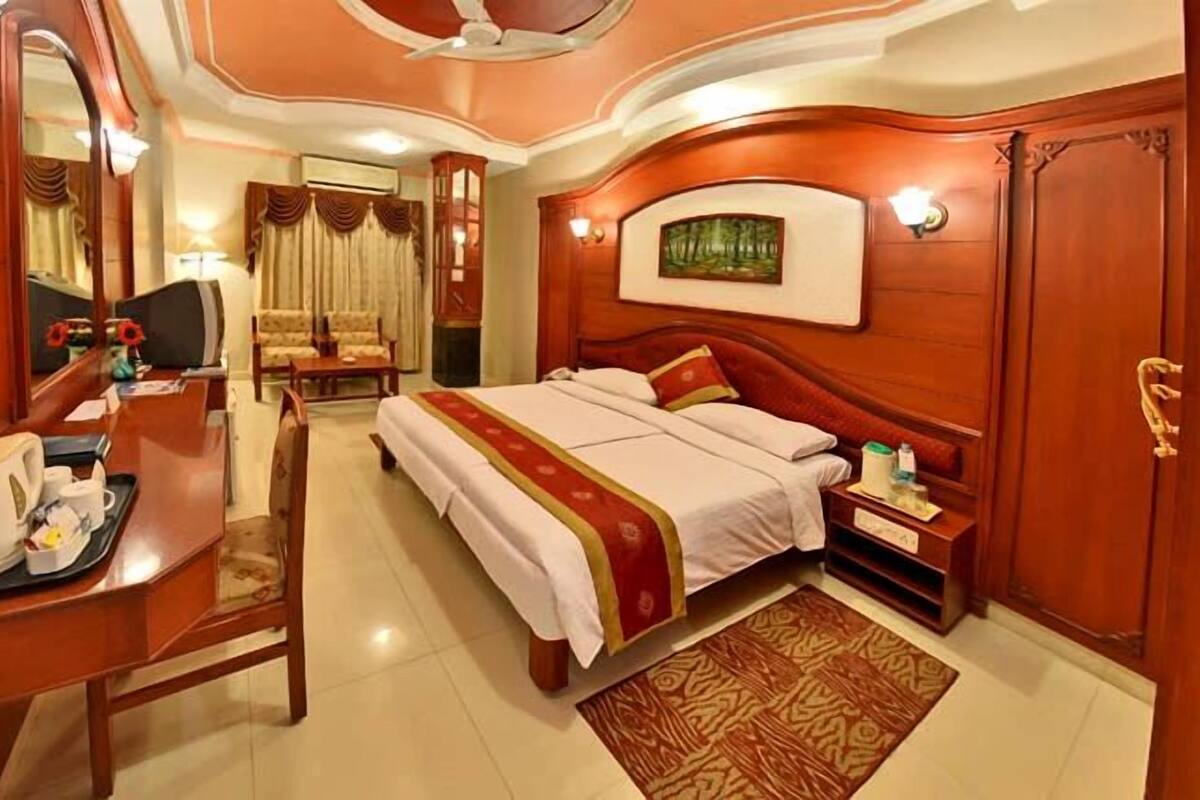 A hotel room with a bed, a desk with a chair, a mirror, a painting on the wall, and a carpet.