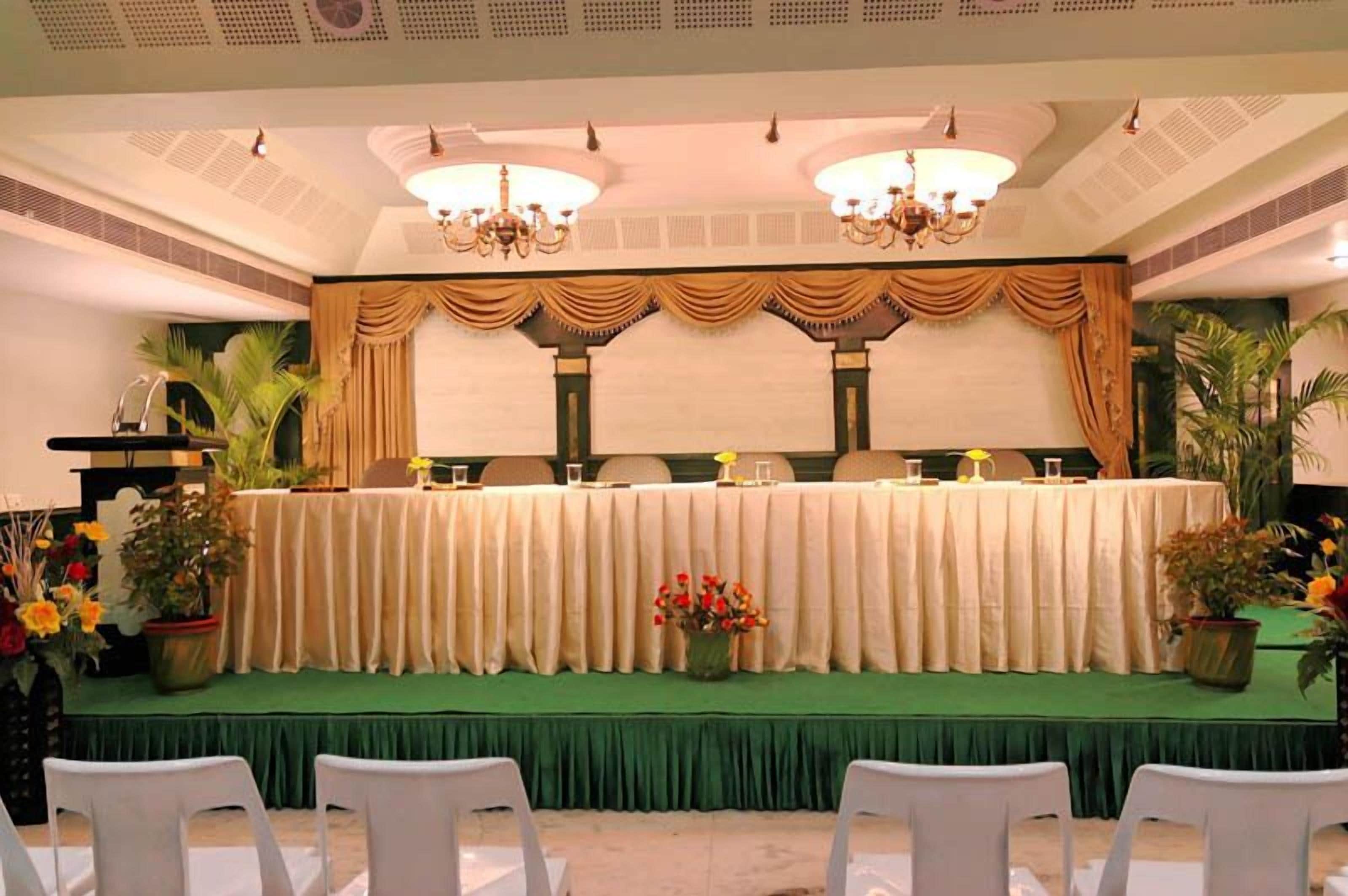 Un salón de banquetes con una larga mesa dispuesta para un evento formal, flanqueado por macetas con plantas y un atril.