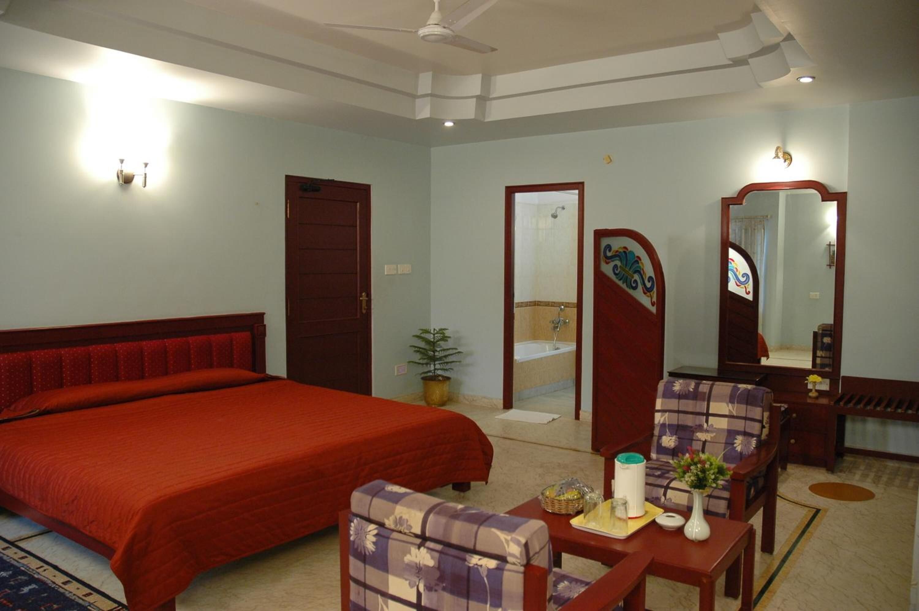 A hotel room with a large bed, a sitting area with a chair and table, a bathroom with a sink and mirror, and a ceiling fan.
