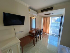 Habitación de hotel con televisor de pantalla plana en la pared, un escritorio de madera con sillas, un armario pequeño y un ventanal con cortinas.