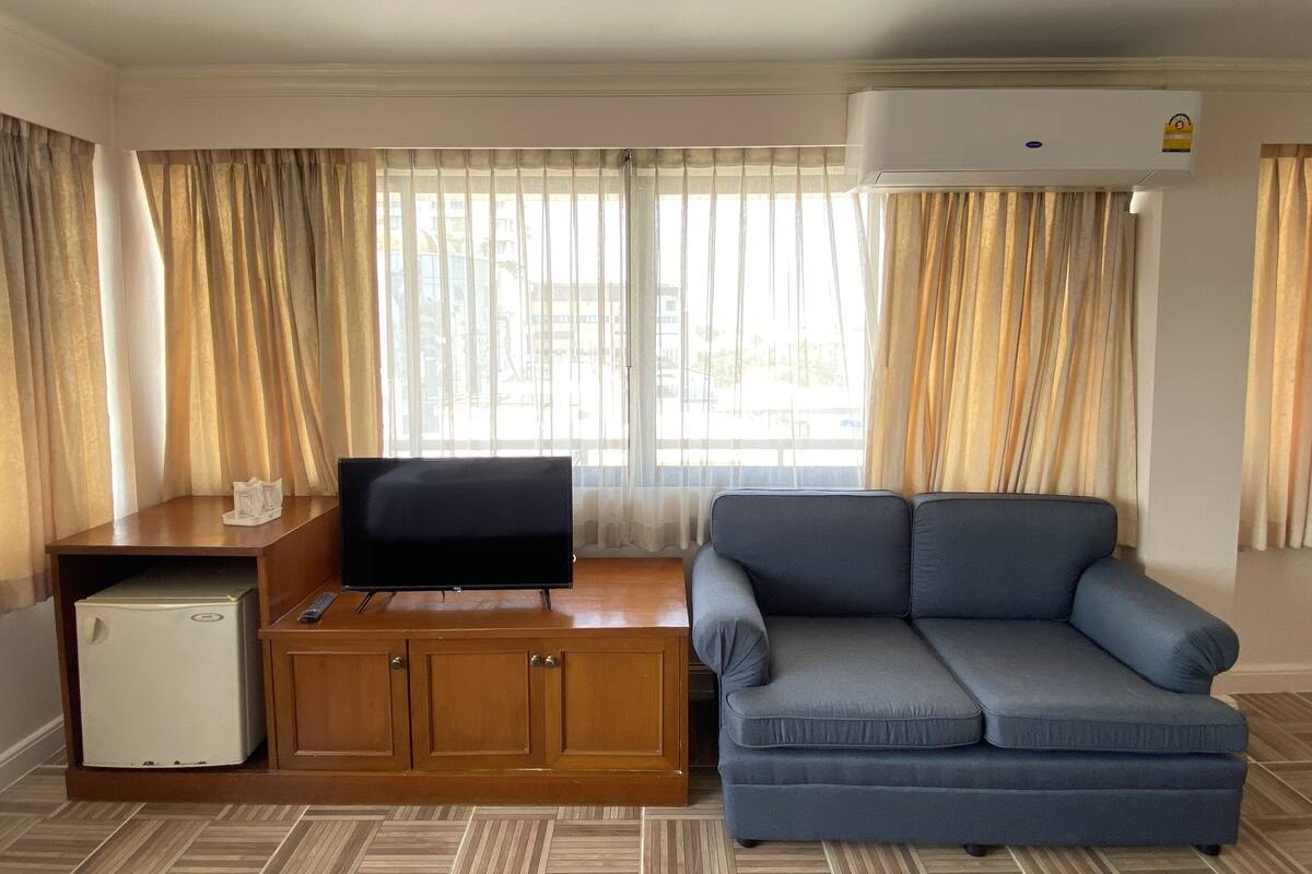 Una sala de estar con sofá, televisor sobre un mueble de madera, un refrigerador pequeño y un ventanal con cortinas.