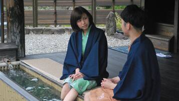 Two individuals seated by a small water feature in a traditional setting with wooden elements and a stone border.