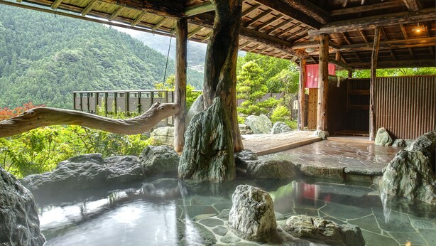 Una estructura de madera tradicional con un baño de aguas termales de piedra, grandes rocas y vistas de vegetación exuberante.