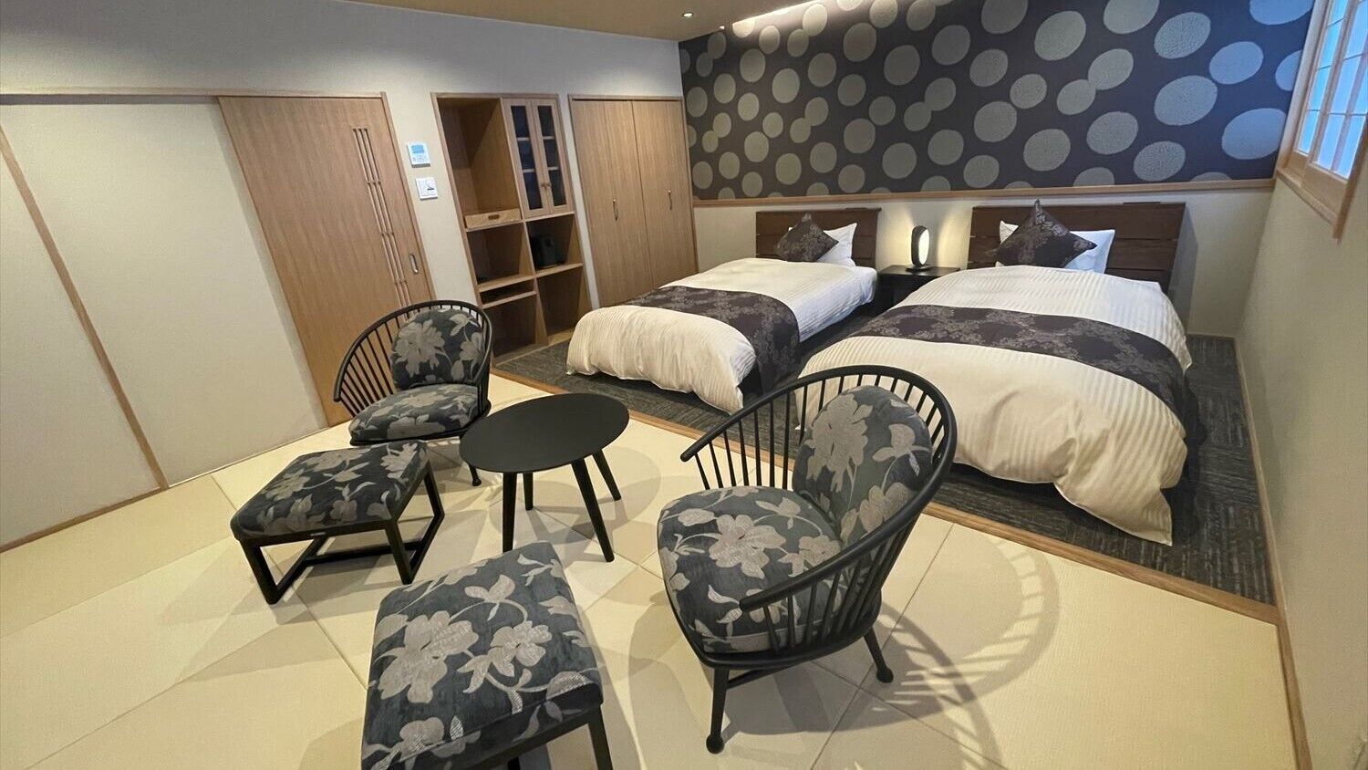 A hotel room with two beds, a seating area with chairs, a wardrobe, and a polka dot wall.