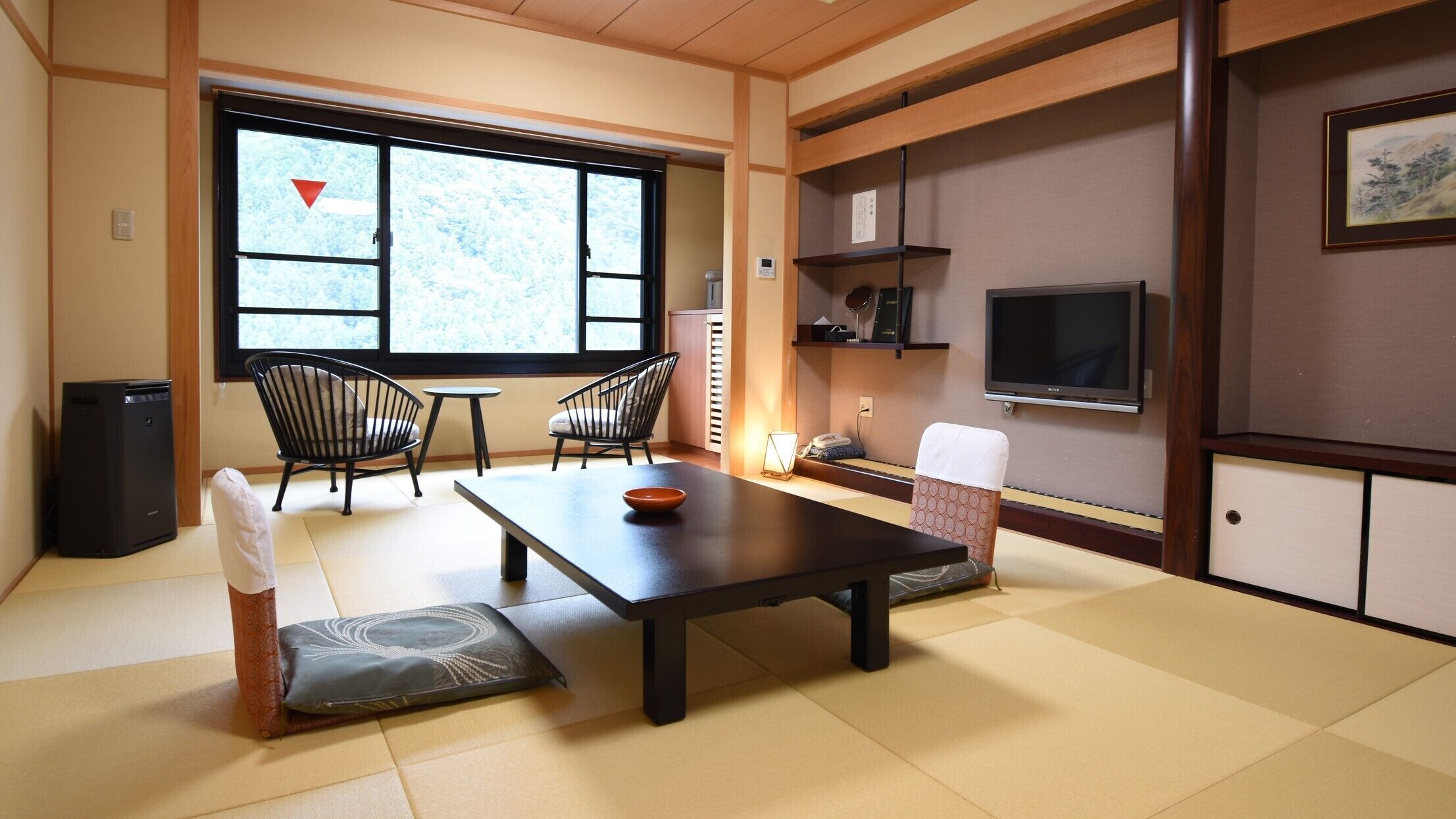 A traditional Japanese-style room with tatami flooring, a low table, two chairs, a TV, and a window with a view of trees.