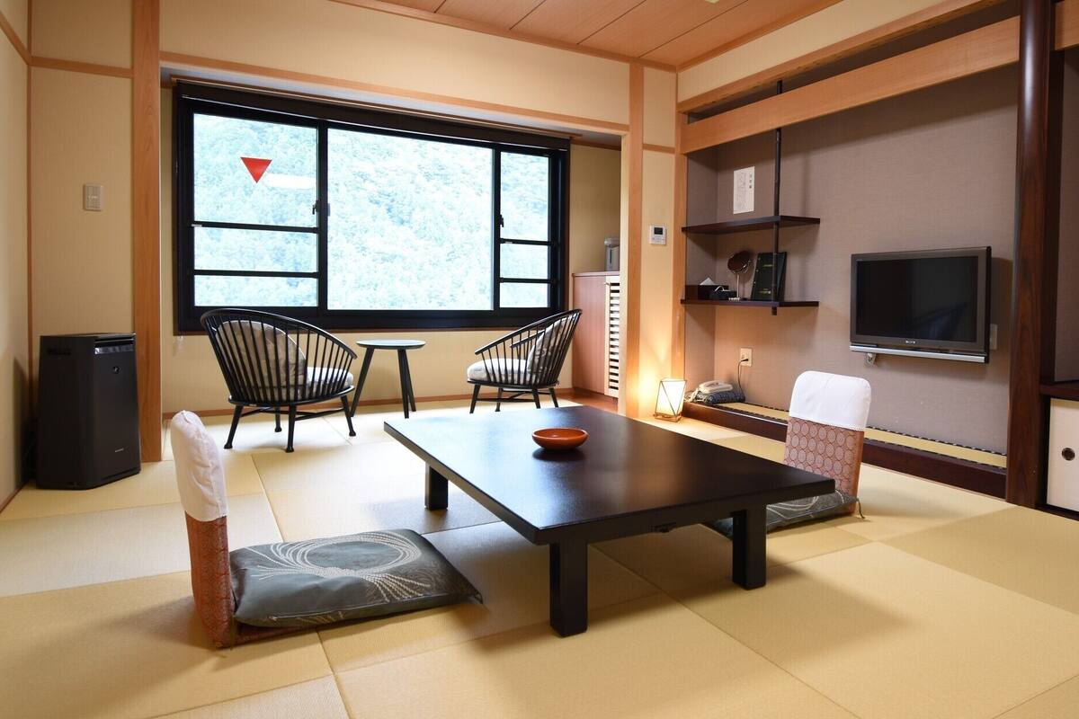 A traditional Japanese-style room with tatami flooring, a low table, two chairs, a TV, and a window with a view of trees.