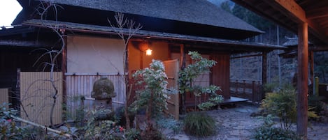 A traditional Japanese house with a thatched roof, a wooden veranda, and a stone pathway leading to the entrance.