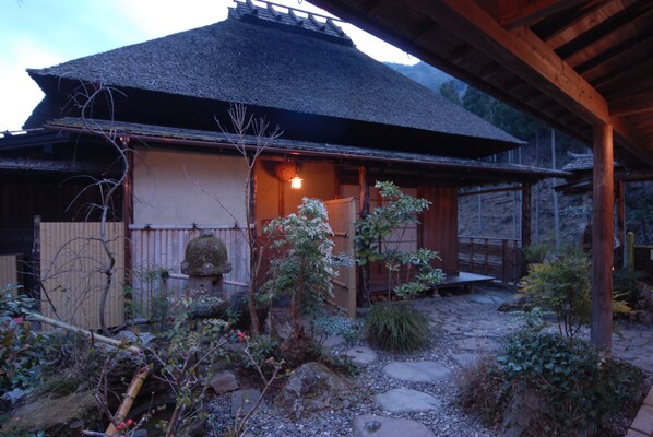 A traditional Japanese house with a thatched roof, a wooden veranda, and a stone pathway leading to the entrance.