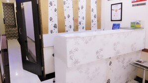 A reception area with a white reception desk, floral patterned wallpaper, framed certificates on the wall, and a clock. - Hotel Aksa (Mumbai)
