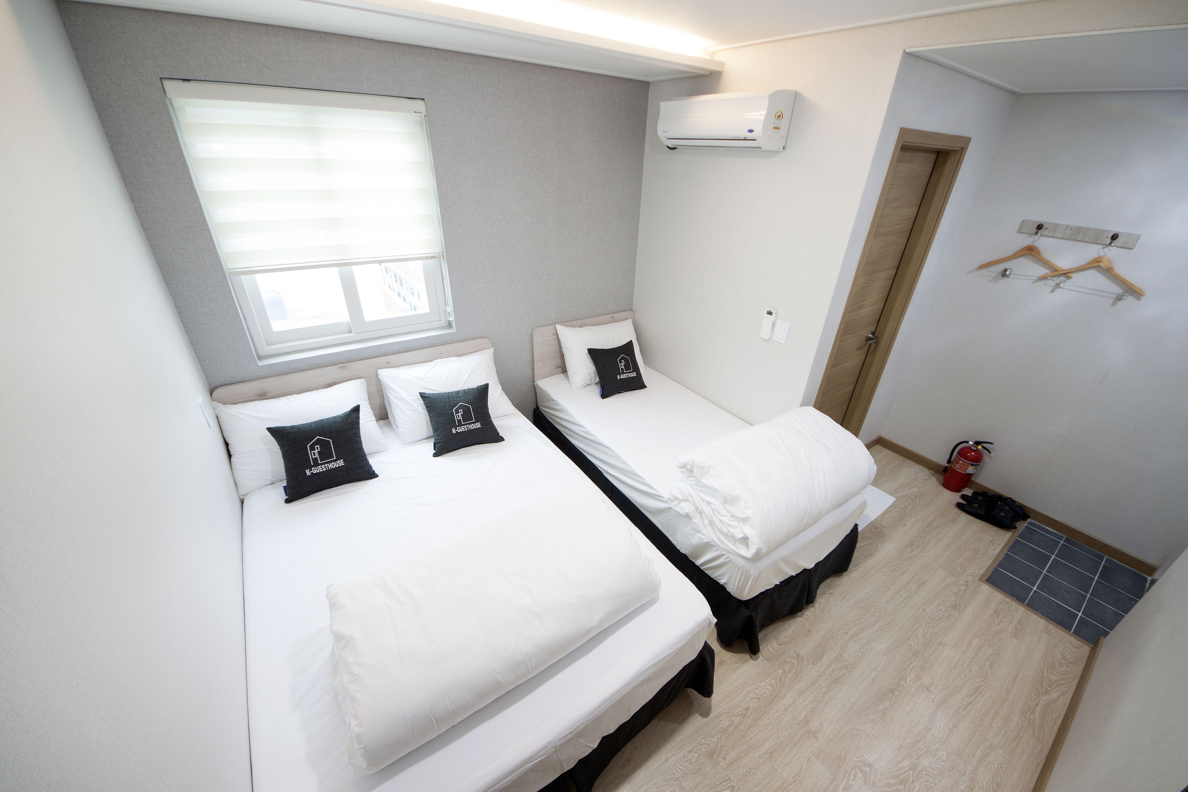 Een kleine hotelkamer met twee bedden, een raam, een airconditioningapparaat, een deur en een brandblusser.