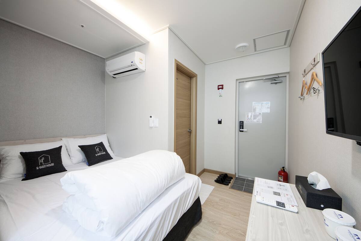 Habitación de hotel con cama, mesita de noche con control remoto, espejo y televisor montado en la pared.