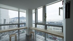 Ein modernes Hotelzimmer mit großen Fenstern und Blick auf die Stadt, einer hölzernen Bank und einem kleinen Hocker.