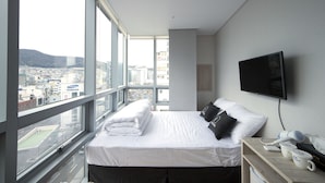 Ein Hotelzimmer mit einem großen Bett, einem Fernseher, einem Fenster mit Blick auf die Stadt und einem Nachttisch aus Holz mit einem Föhn und einer Tasse.