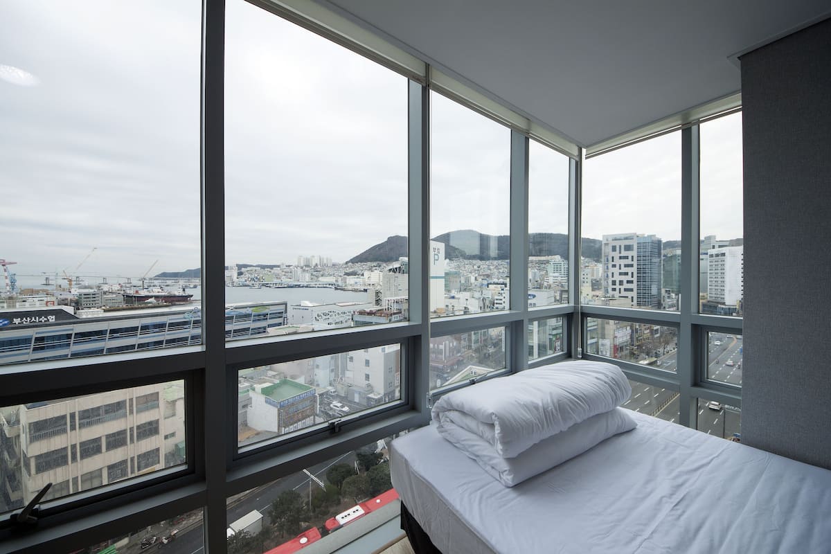 Habitación de hotel con un ventanal grande que ofrece vistas a la ciudad, una cama con ropa blanca y vistas a un puente y edificios.