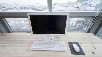 A computer desk with a monitor, keyboard, and mouse, positioned near a large window overlooking a cityscape.