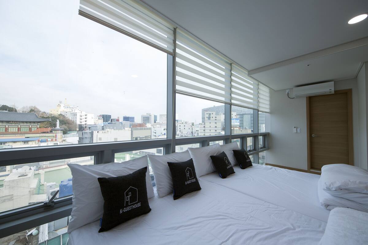 Habitación de hotel con una cama grande, varios almohadones, una puerta de madera y vistas a la ciudad a través de amplios ventanales.