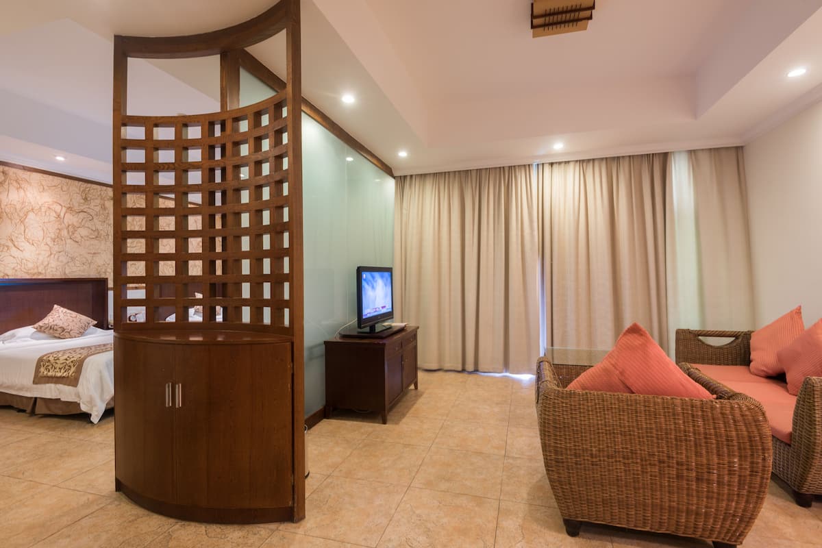 Une chambre d’hôtel comprenant un lit, une cloison en bois, un téléviseur posé sur un meuble, un canapé en osier avec des coussins et une grande fenêtre avec des rideaux.