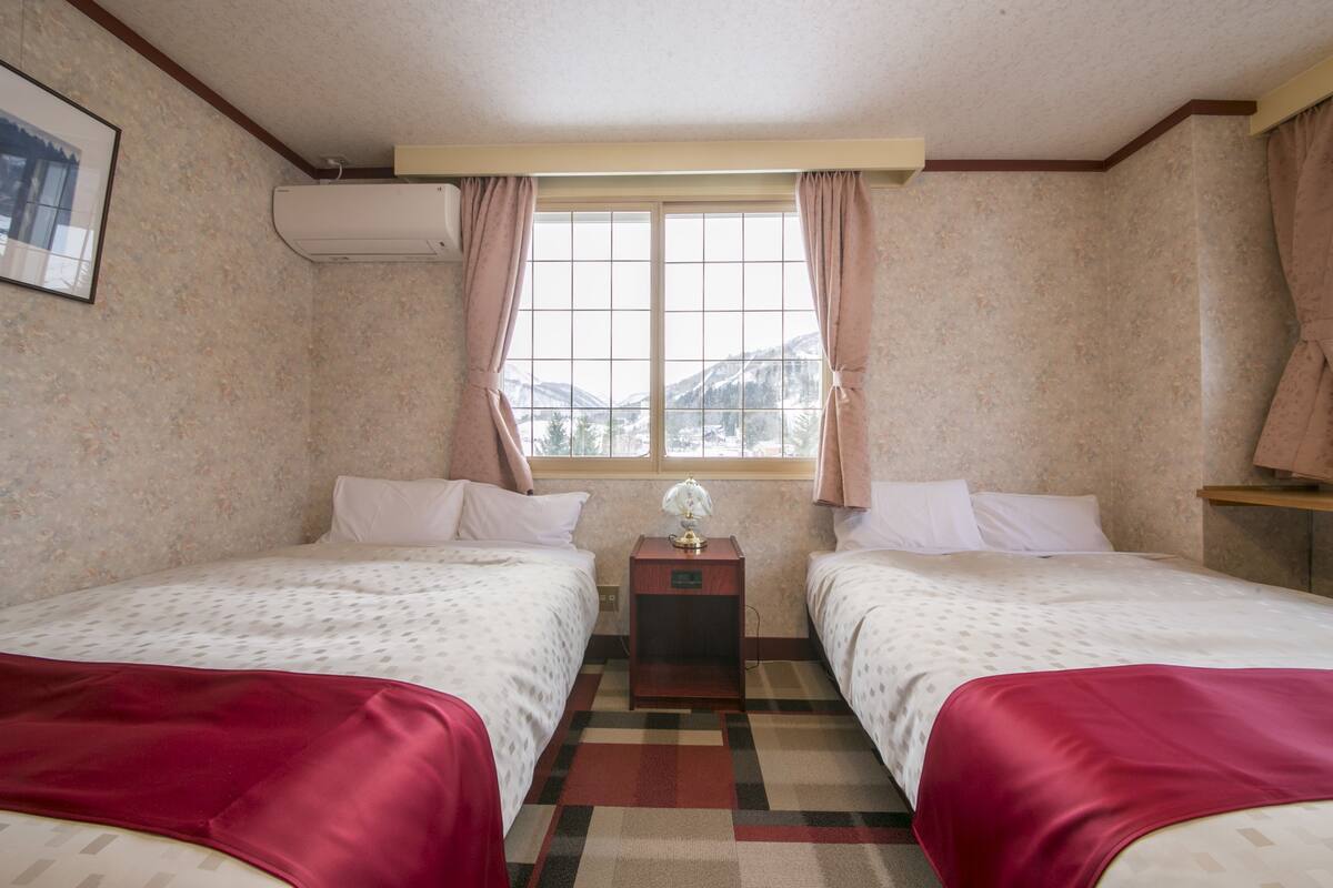 A hotel room with two beds, a nightstand with a lamp, a window with curtains, and a view of mountains.