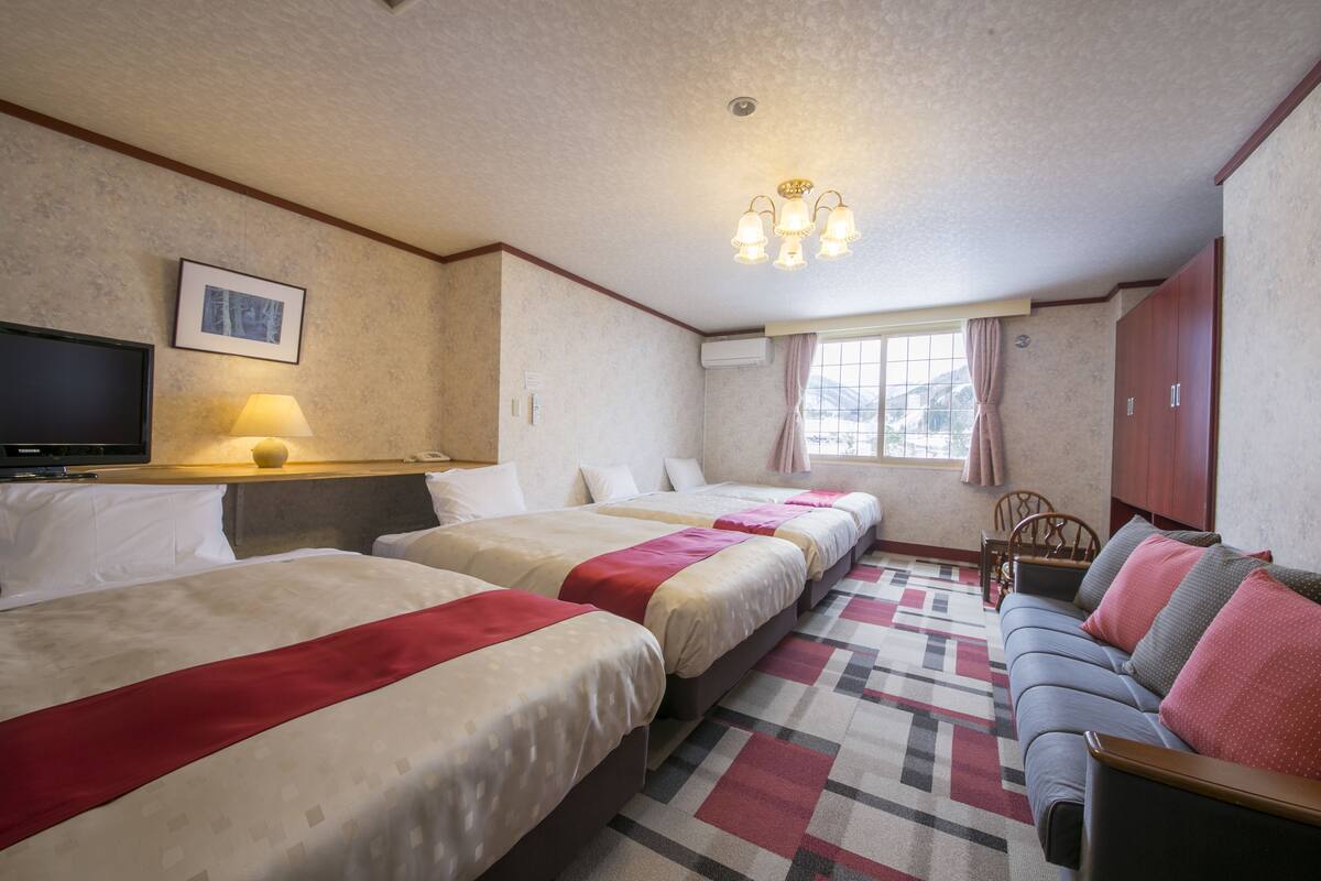 A hotel room with three beds, a sofa, a TV, and a window with curtains.