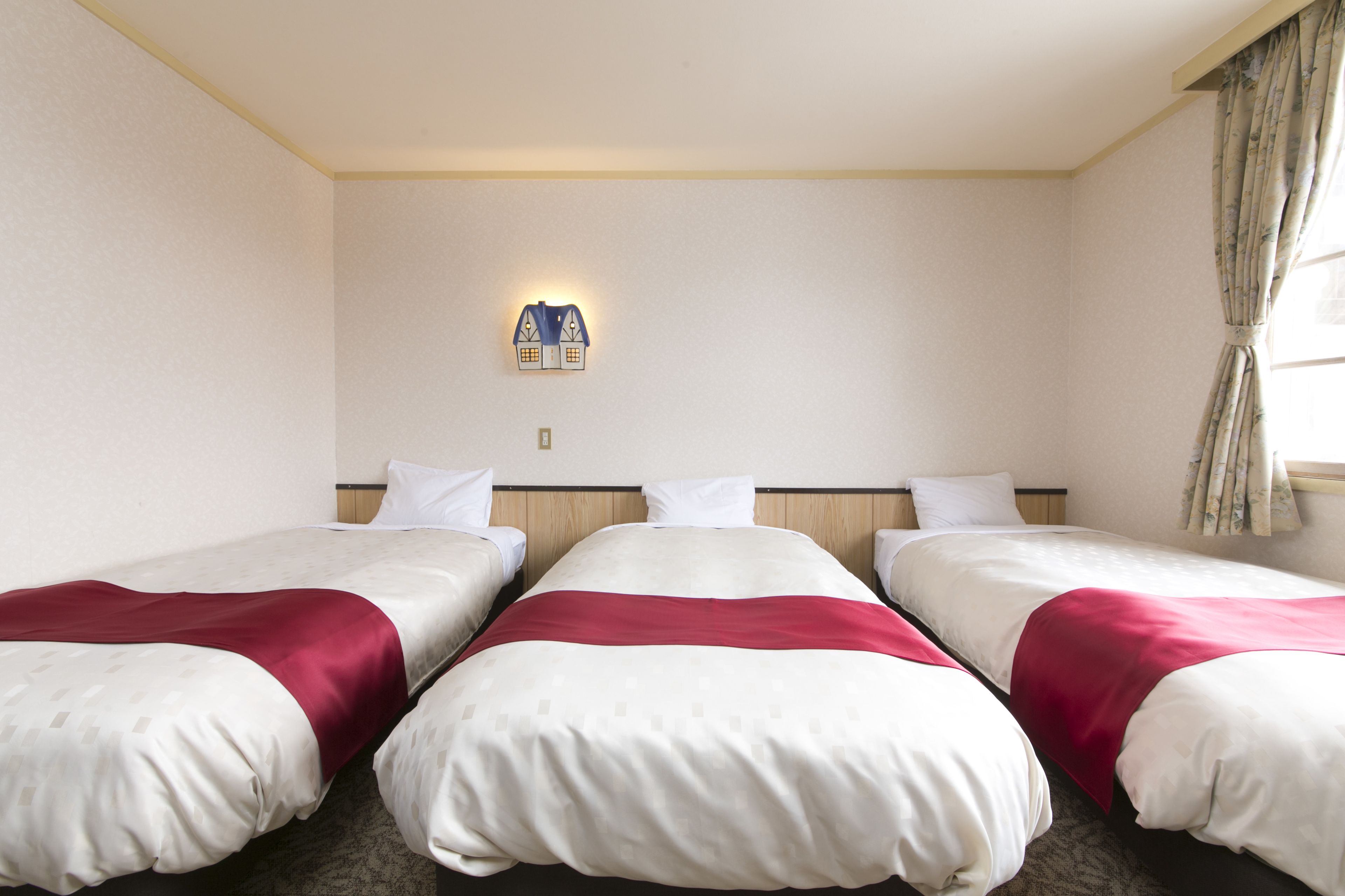 Une chambre d’hôtel avec trois lits simples, un luminaire fixé au mur et une fenêtre avec des rideaux.