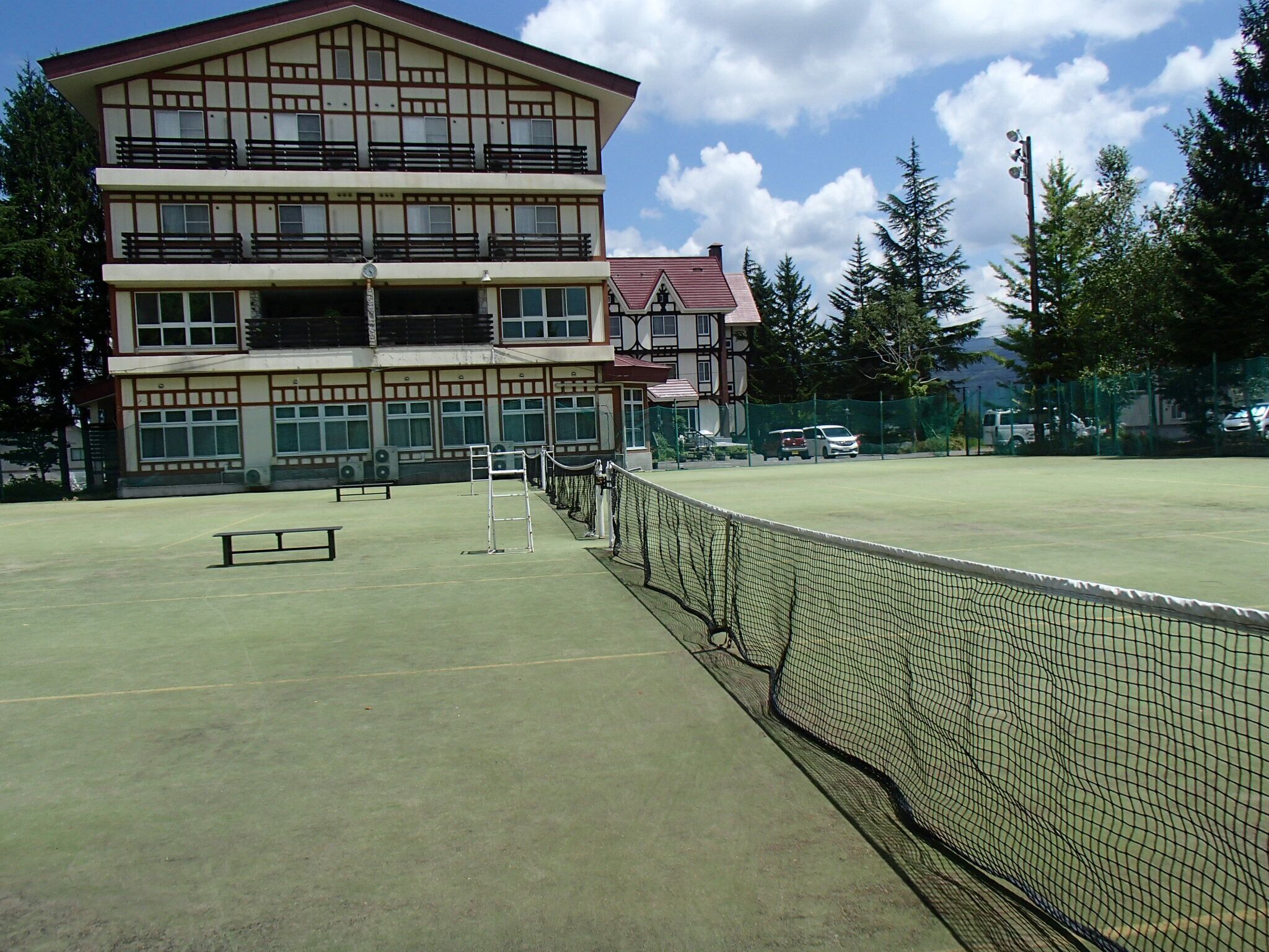 Un court de tennis avec un filet, un bâtiment doté de nombreuses fenêtres et de balcons, et un espace de stationnement avec des voitures.