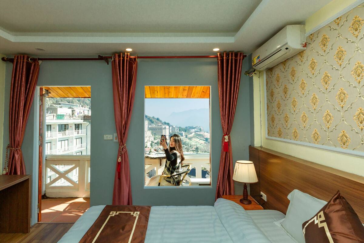 Ein Hotelzimmer mit Bett, einem fenster mit Vorhängen, Blick auf eine Stadtlandschaft und einer Wand mit einem Musterdesign.