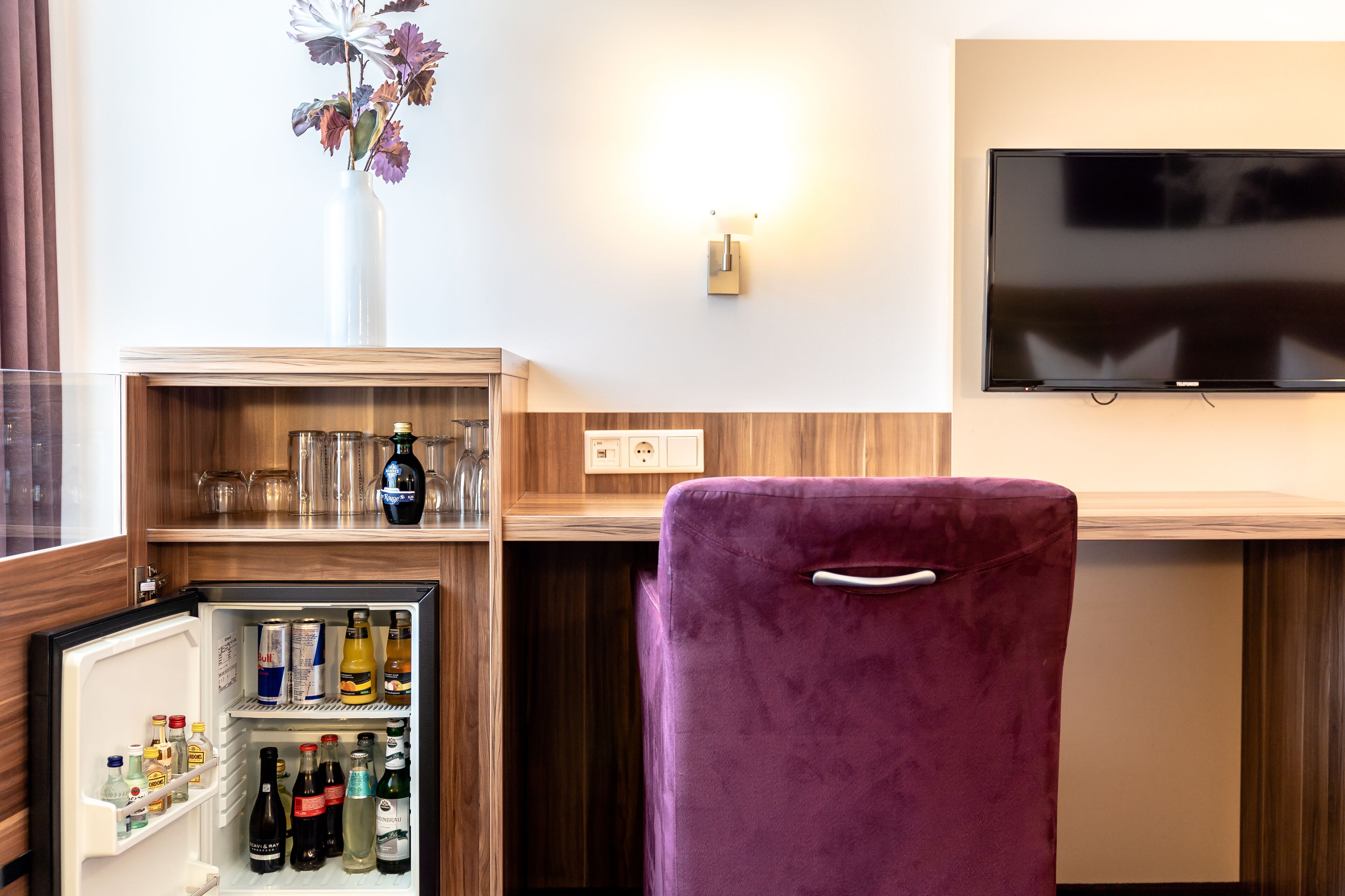 A compact room with a purple chair, a small fridge stocked with beverages, a wooden shelf with glassware, and a mounted TV.