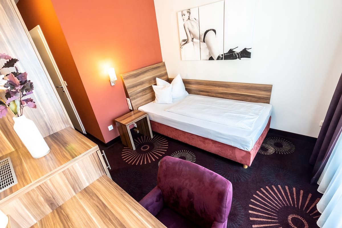 A hotel room with a wooden headboard, a bed with white linens, a purple armchair, and a wall-mounted artwork.