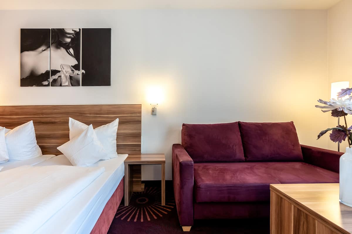 A hotel room with a bed, a wooden headboard, a red sofa, a small table, and a wall-mounted light.