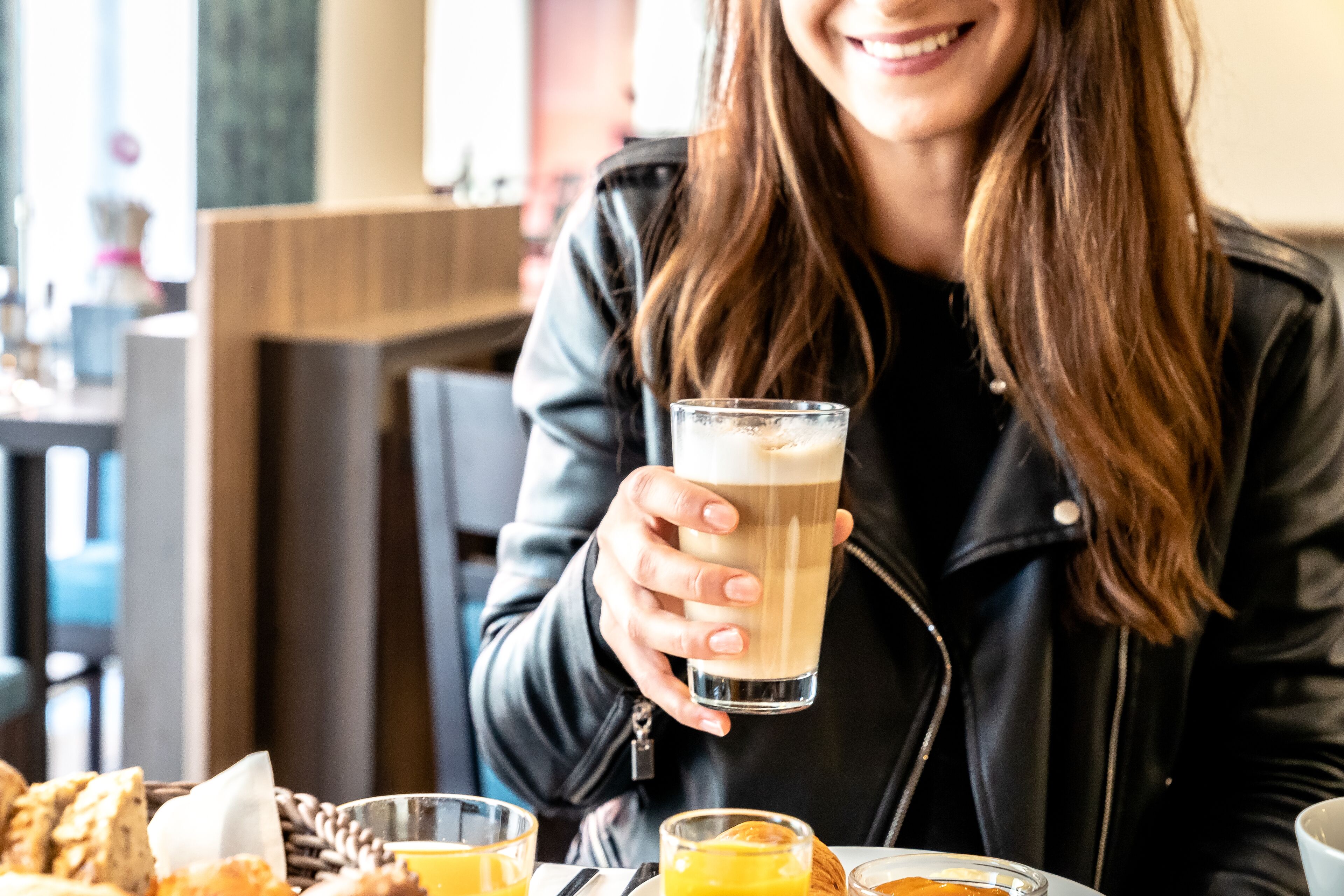 Una persona sosteniendo un vaso de una bebida clara, posiblemente café o té, en una mesa con una cesta de pan y un vaso de jugo de naranja.