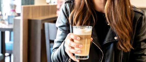 Una persona sosteniendo un vaso de una bebida clara, posiblemente café o té, en una mesa con una cesta de pan y un vaso de jugo de naranja.