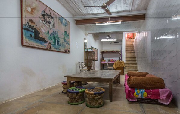 Una habitación con una mesa de madera, taburetes de mimbre, un sofá con cojines, un ventilador de techo y una escalera que lleva a otra zona.