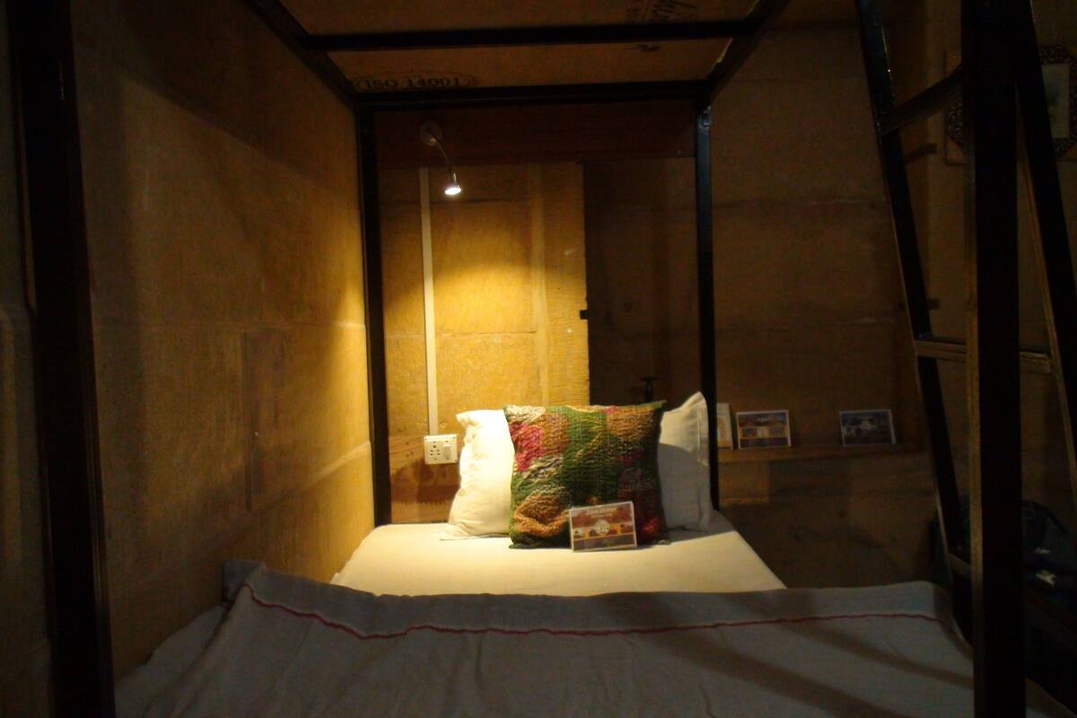 A narrow, dimly lit room with a single bed, a quilt, and a wall-mounted light fixture.