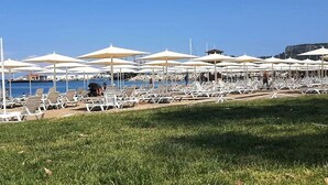 Een strand met rijen ligbedden en parasols, een zwembad op de achtergrond, en een gazon in de voorgrond.