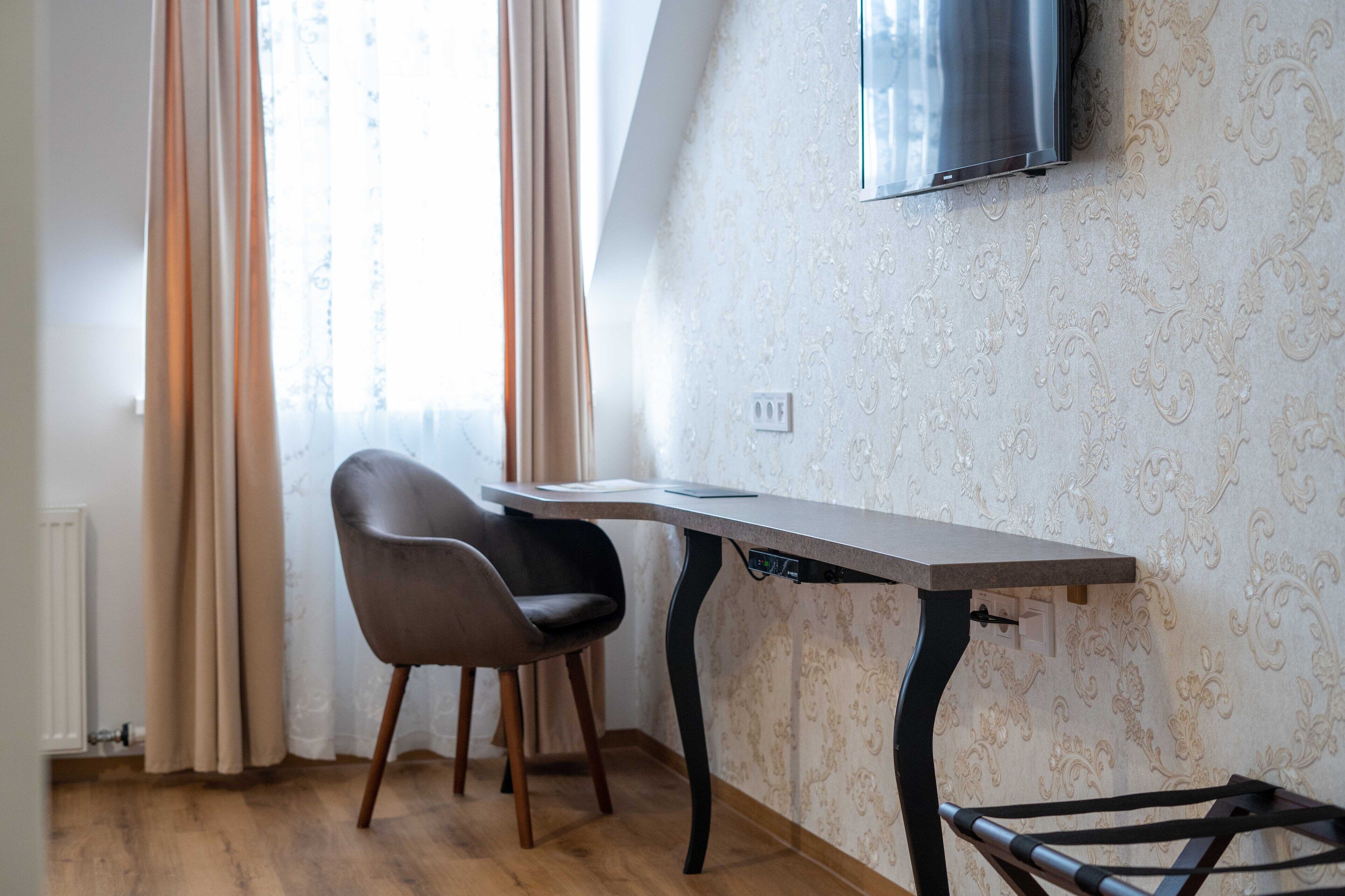 Een kamer met een houten vloer, een bureau met stoel, een aan de muur bevestigde televisie en een raam met gordijnen.