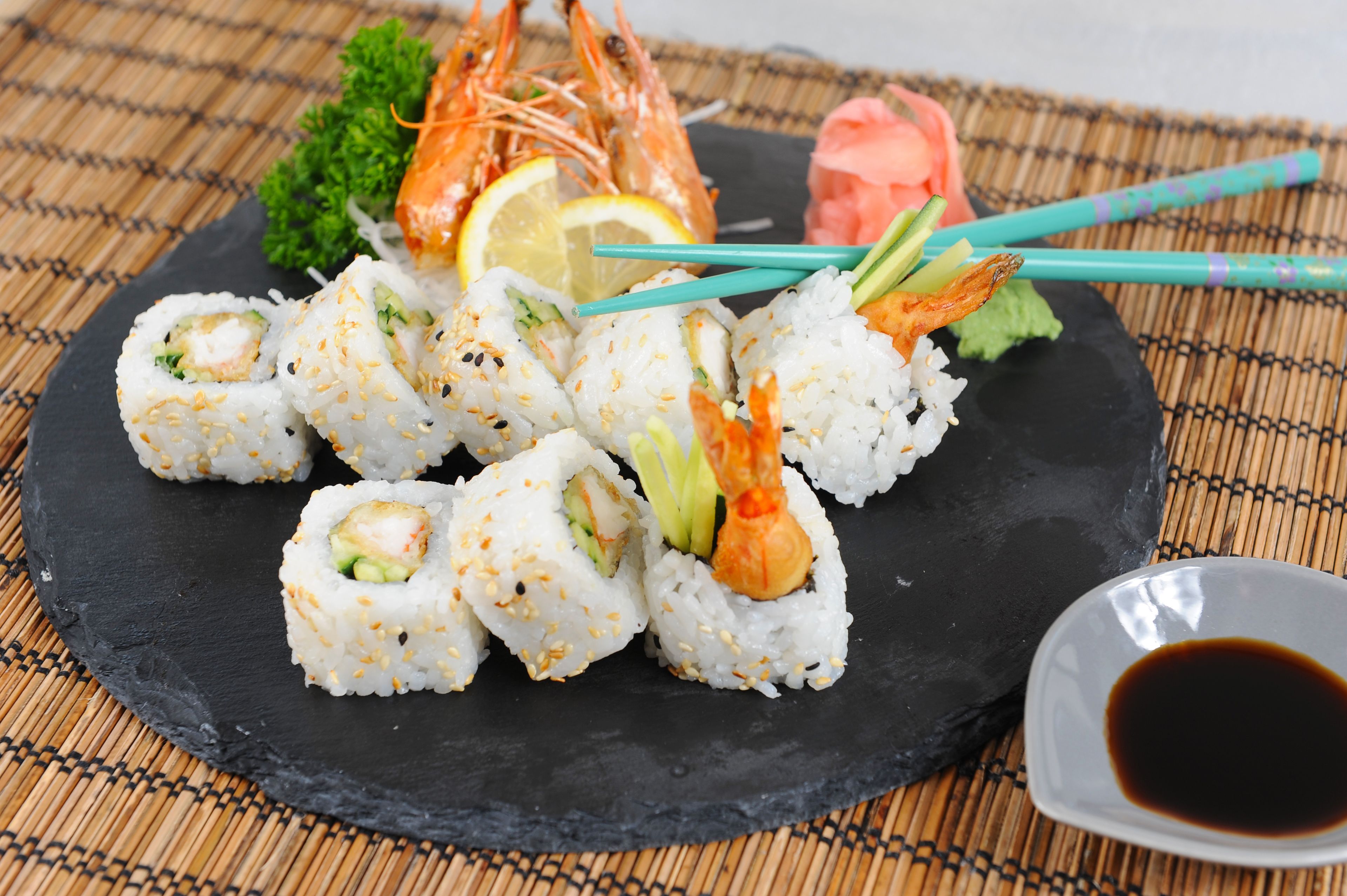 A black slate plate with sushi rolls, shrimp, lemon, and a small dish of soy sauce.