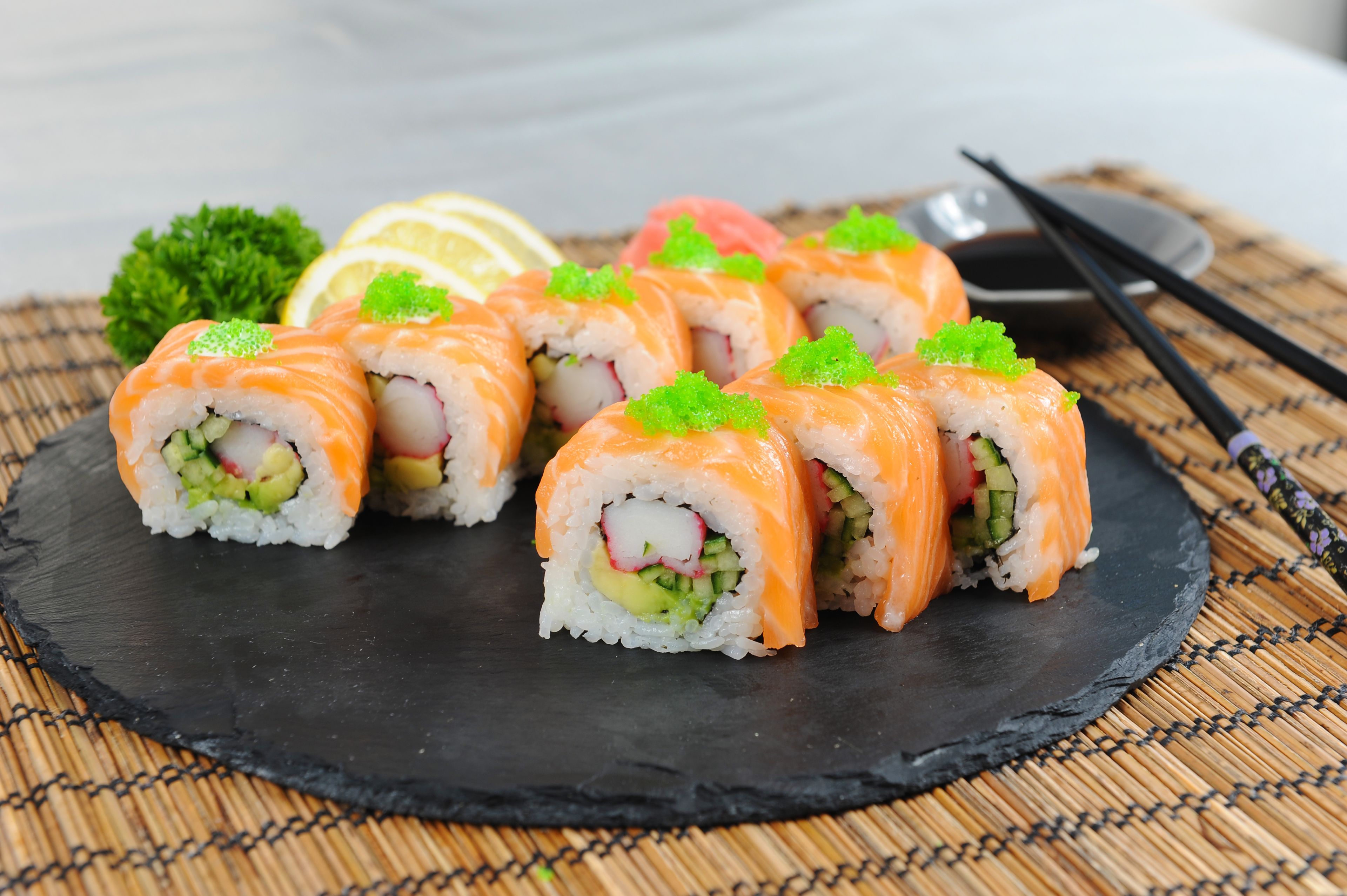 A black slate plate with six pieces of salmon sushi, garnished with green caviar, on a bamboo mat.